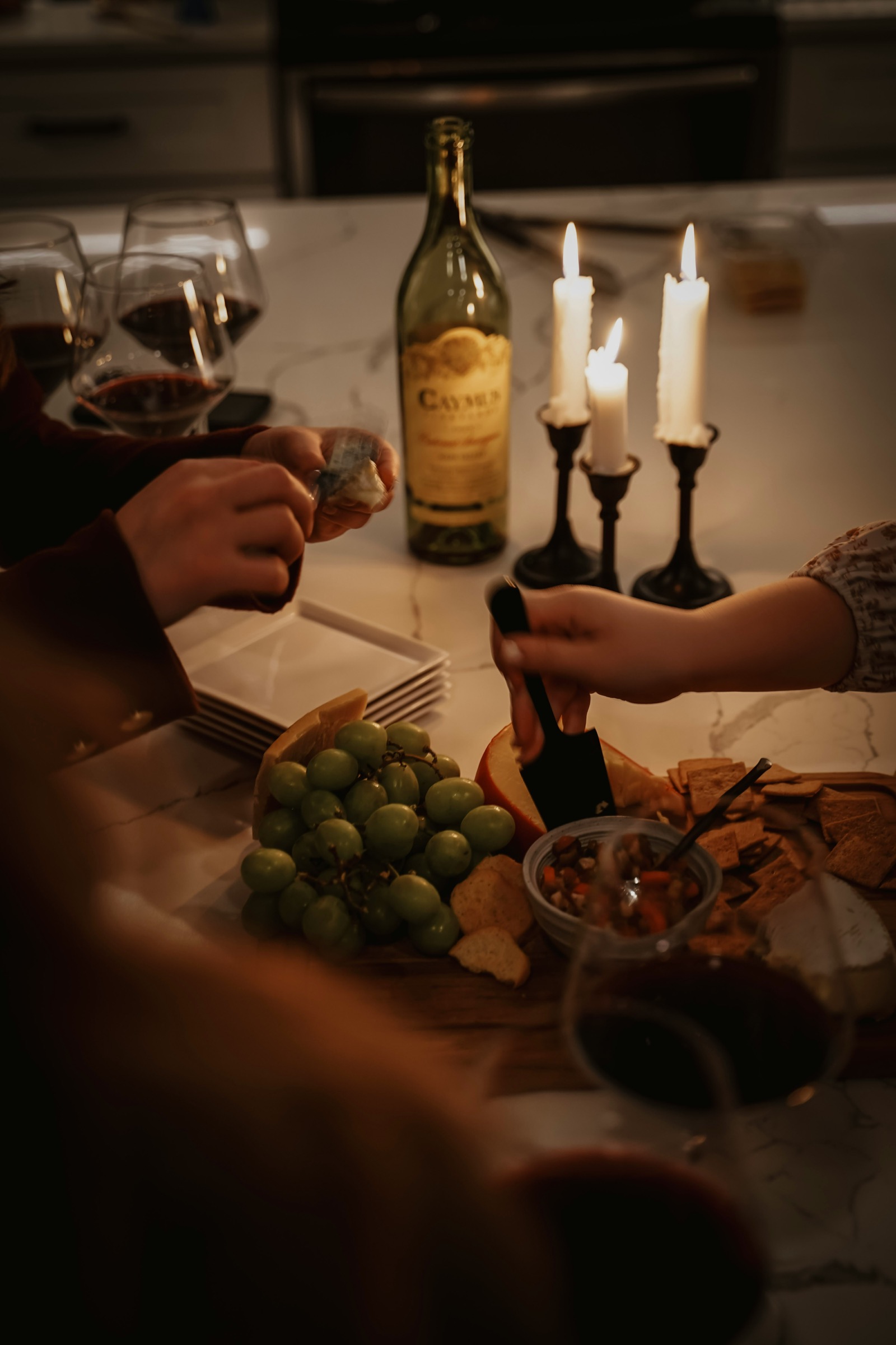 Candlelit dinner table with wine glasses, hands reaching for food, warm intimate atmosphere